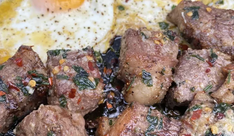 Ribeye steak bites covered in garlic-infused butter, served with sunny-side-up eggs on a plate.