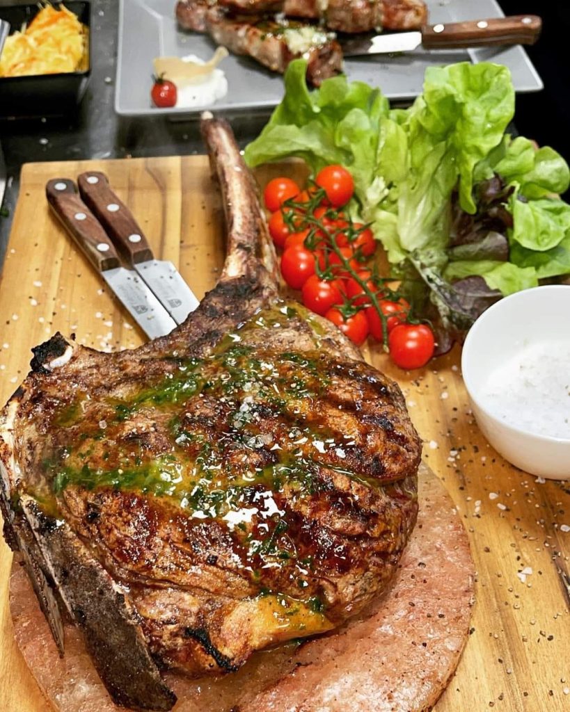thick-cut Tomahawk steak seasoned with herbs and spices, sizzling on a hot grill with flames leaping up around it.
