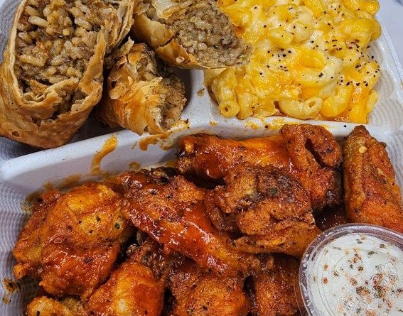 A platter piled high with Cajun dishes: Dirty Rice Boudin Egg Rolls (golden brown and crispy), Crack Mac and Cheese (creamy and bubbly), and Lemon Bae Wings (glazed and glistening).