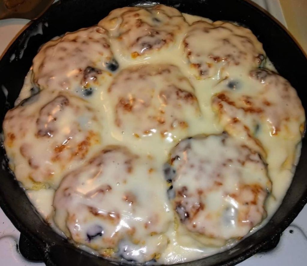 A close-up photo of golden brown sweet blueberry biscuits on a baking sheet. Fresh blueberries are visible throughout the fluffy biscuits.